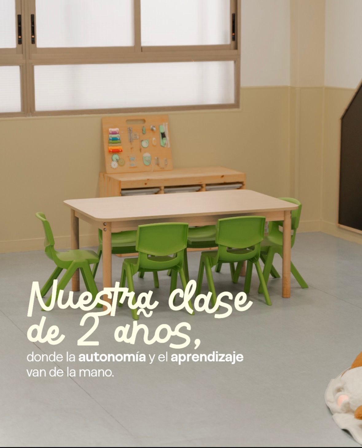 Aula de dos años con mesa baja, sillas verdes, un panel de actividades sobre el mueble y una pizarra. Texto en la imagen: nuestra clase de 2 años, donde la autonomía y el aprendizaje van de la mano.