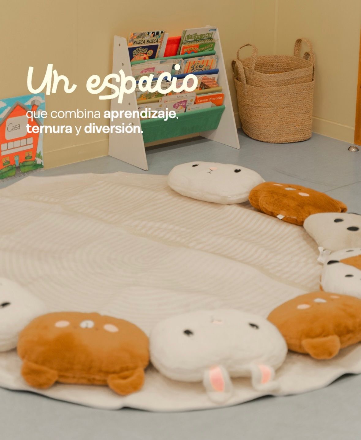 Rincón de lectura con un expositor de cuentos infantiles, un cesto de fibra natural y una alfombra rodeada de cojines con forma de animales. Texto en la imagen: un espacio que combina aprendizaje, ternura y diversión.