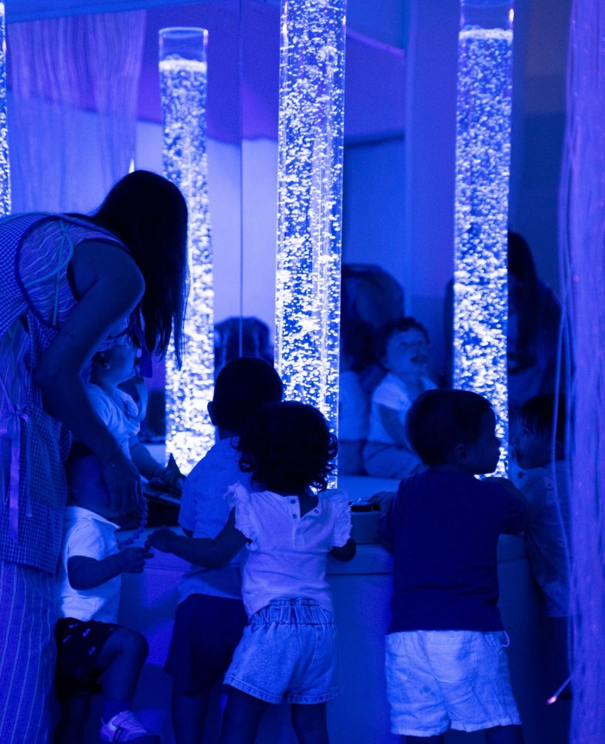 Varios niños y su tutora frente a las columnas de burbujas iluminadas en azul, reflejados en los espejos de la sala.